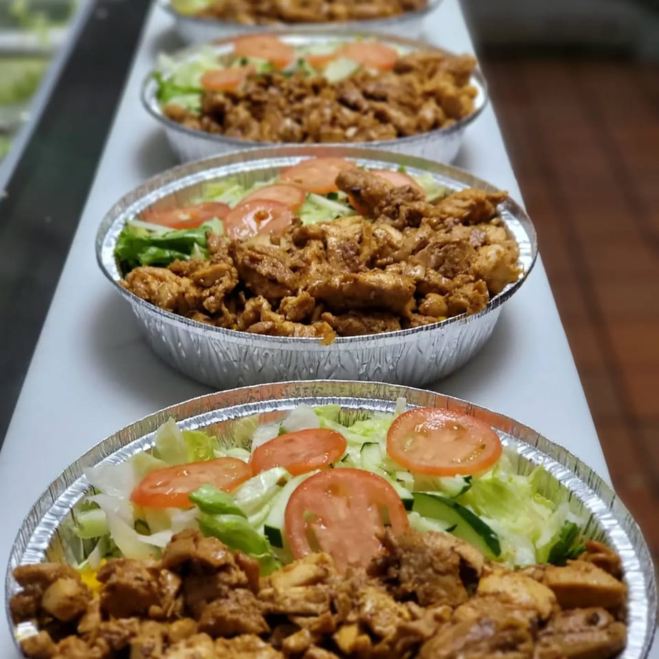 Multiple foil containers of halal chicken over rice with lettuce, tomatoes, and cucumbers.