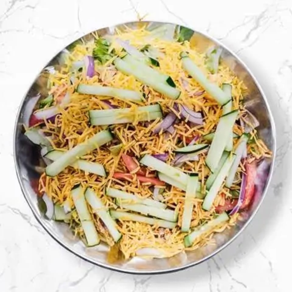 A close-up of a fresh salad with shredded cheese, sliced cucumbers, red onions, and tomatoes.