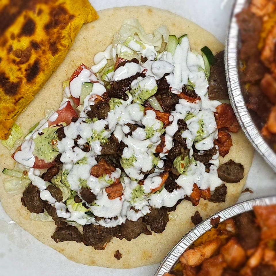 Halal platter with meat, lettuce, tomatoes, cucumbers, and white and green sauces on pita bread.